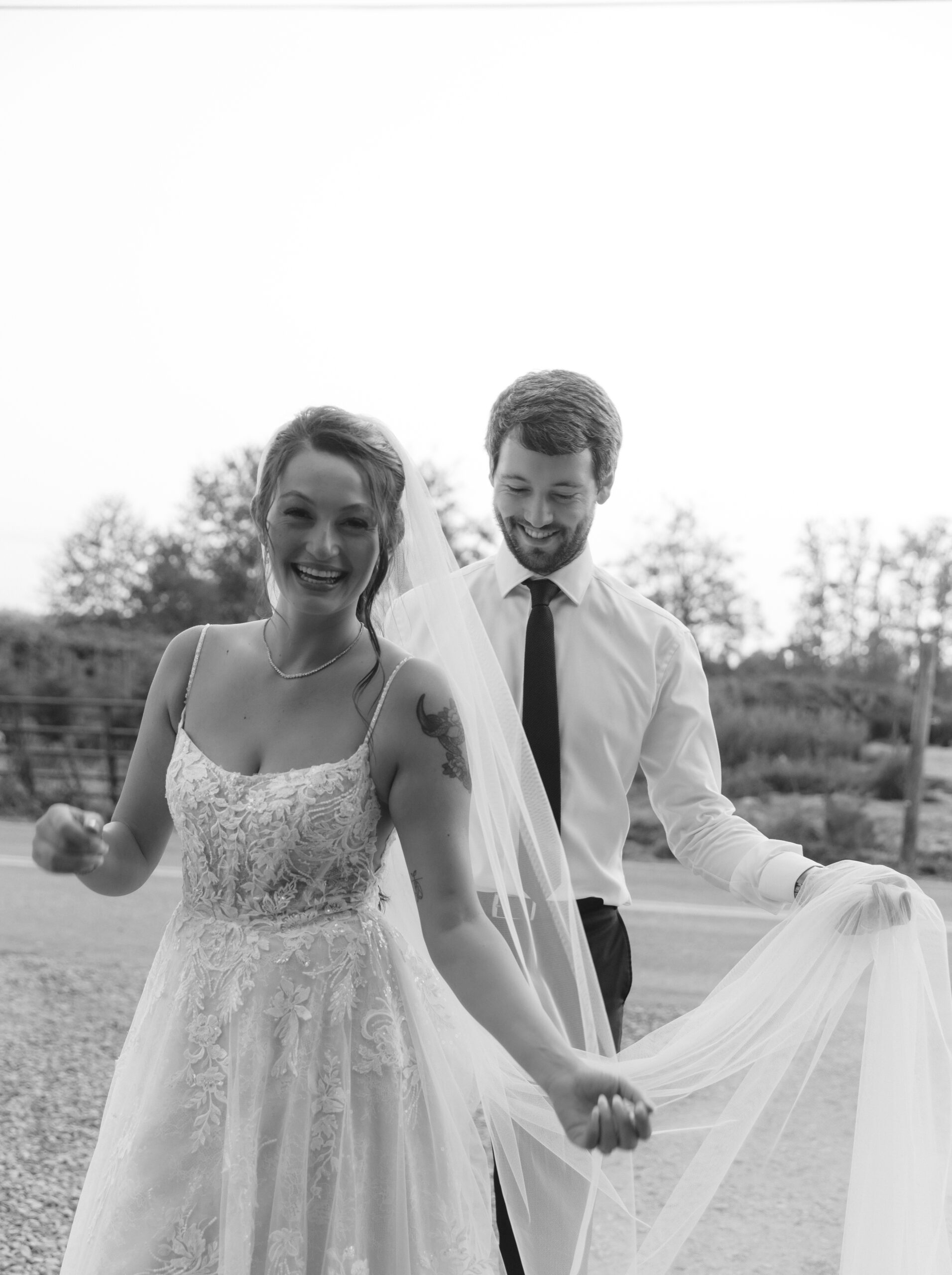 Black and white photo of bride smiling as groom adjusts her veil during golden hour after the ceremony.